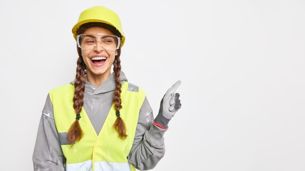 Positive woman builder on construction site demonstrates something over blank space wears hard hat protective glasses uniform has engineering career isolated over white wall. Safety apparel