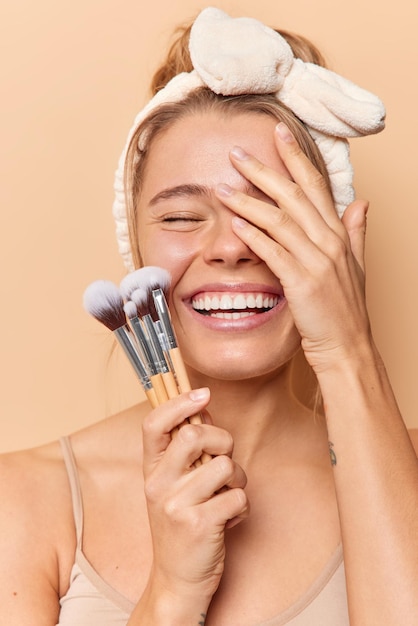 Positive woman covers face with hand keeps eyes closed smiles broadly wears headband holds cosmetic brushes applies makeup has well cared skin isolated over beige background. Beauty concept.