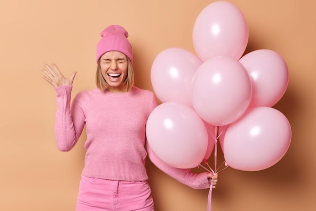 Positive woman enjoys house party moves in new apartment keeps palms raised and exclaims gladfully wears pink clothes poses with inflated balloons isolated over beige background Celebration