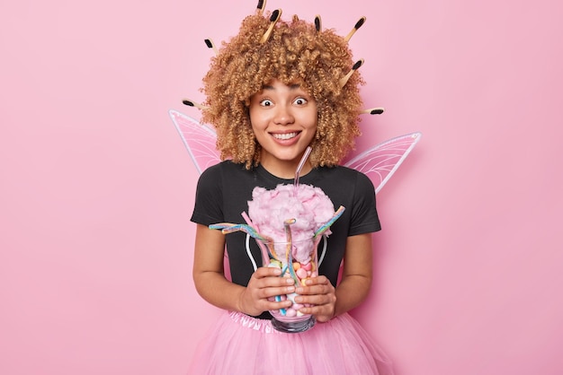 Positive woman fairy holds glass of delicious sweet desserts\
pretends being magical character wears black t shirt pleated skirt\
and wings poses against pink background tooth fairy with\
sweets