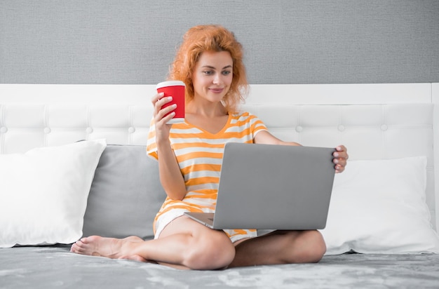 집에서 노트북으로 커피를 마시는 긍정적인 여성 프리랜서 프리랜서 여성이 집에서 일함 프리랜서 침대에서 노트북을 들고 일하는 여성