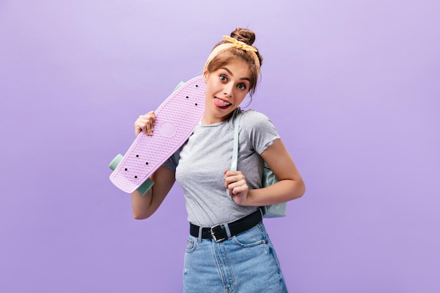 Positive woman in grey shirt shows tongue and holds longboard Happy girl with yellow summer headband posing on isolated background