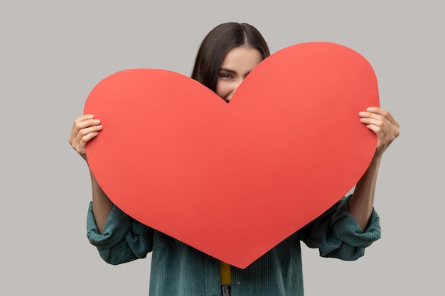 Positive woman hiding face behind big red heart and looking at camera feeling affection fondness