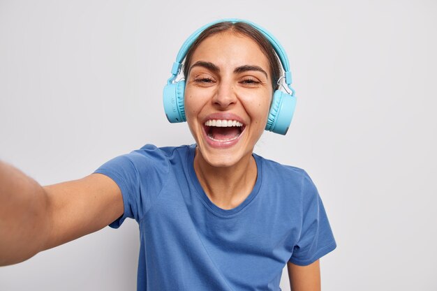 Positive woman listens music in wireless headphones dressed casually smiles broadly makes photo of herself poses over white wall