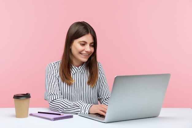 Позитивная женщина-офисный работник в полосатой рубашке сидит на рабочем месте с улыбкой, пьет кофе и делает заметки, работая на ноутбуке, хорошее настроение. Внутренний студийный снимок на розовом фоне.