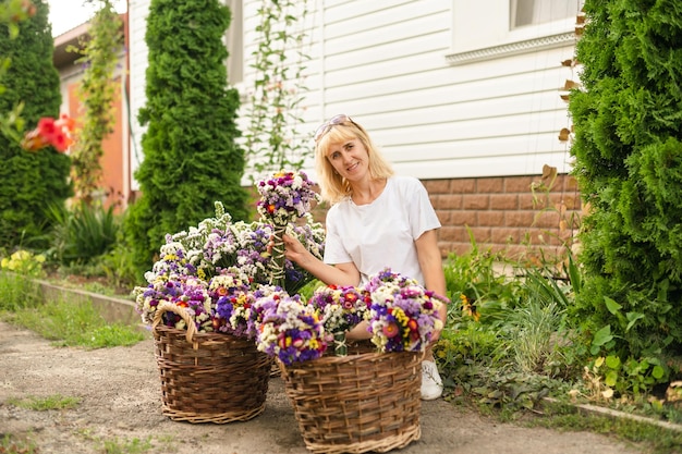 Positive woman sitting in the yard at the cottage with baskets of flowers and posing for the camera with a smile on his faceBlonde woman sitting near the house with baskets of beautiful dried flowers