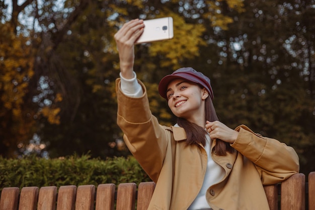 公園で楽しみながら携帯電話で自画像を撮るトレンチとキャップの肯定的な女性