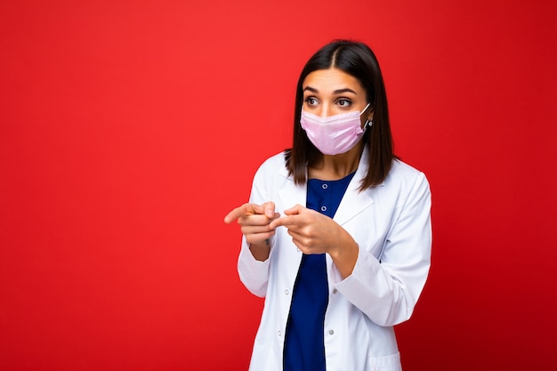 Positive woman in virus protective mask on face against coronavirus and white medical coat isolated on the background and pointing finger at copy space