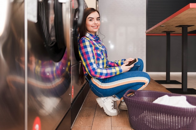 Positive woman waiting for the end of the laundry while sitting on the floor in a laundromat