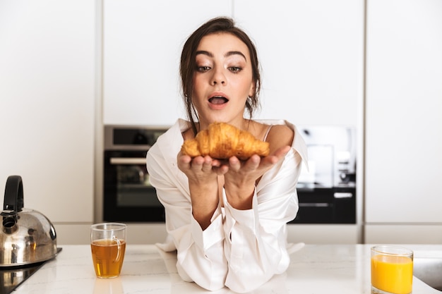 Donna positiva che indossa abiti di seta che mangia croissant, durante la colazione in cucina a casa