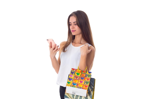 Positive woman in white blouse and skirt with sunglasses holding shopping bags and using smartphone