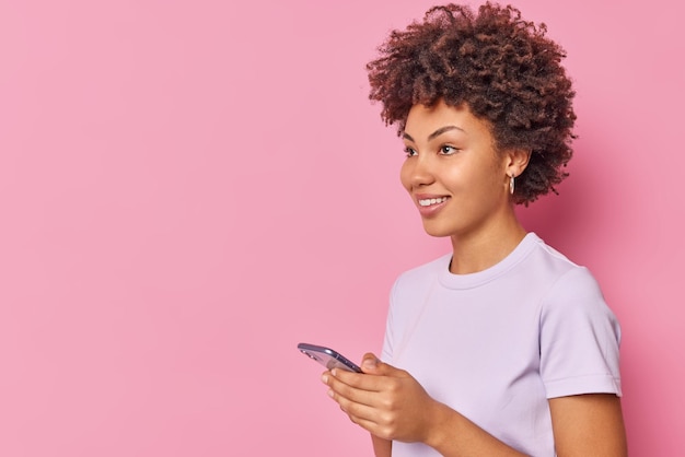 Positive woman with curly hair uses mobile phone chats inn social networks sends text messages reads positive feedback under her post wears casual t shirt isolated over pink wall