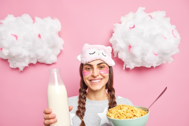 Positive woman with two pigtails going to have breakfast eats cereals with milk smiles gently dressed in nightwear