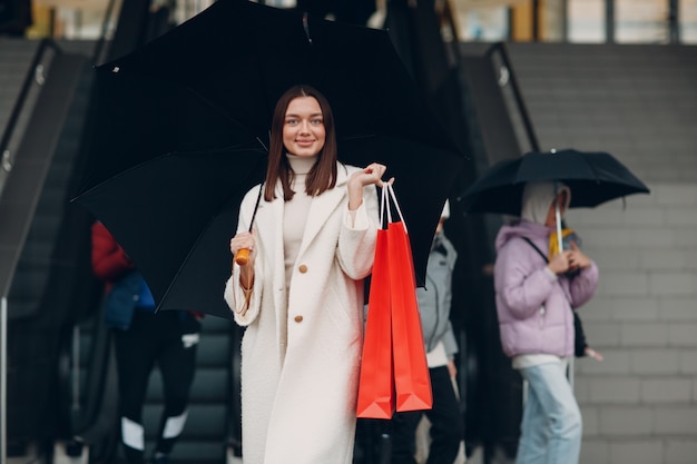 Positivo giovane donna adulta che porta borse della spesa di carta e ombrello in mani