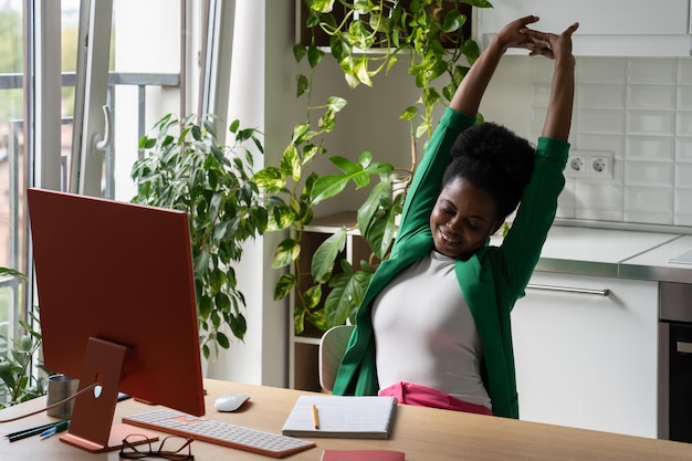 Photo positive young african american woman millenial office worker at table with computer and warming up