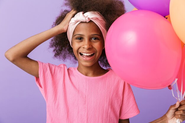 positive young african girl kid posing with balloons isolated over purple wall,.