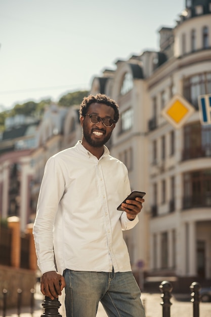 Positive young Afro American man on sunny day standing with smartphone in the street and smiling