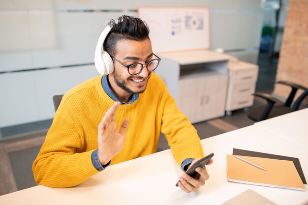 Positivo giovane specialista di affari arabi in occhiali seduto alla scrivania e saluto il collega pur avendo una comunicazione faccia a faccia tramite app mobile