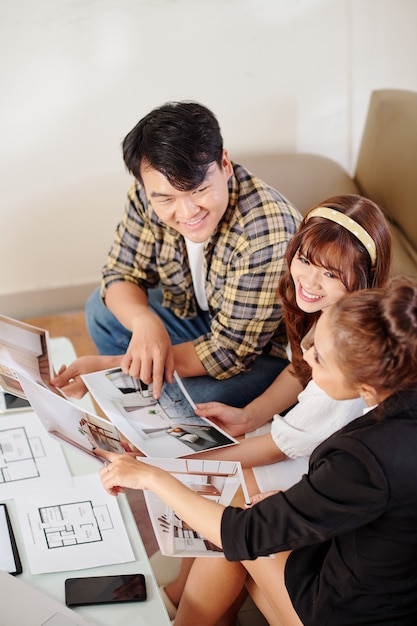 Positive young Asian couple having meeting with real estate manager and discussing photos and layouts of aparments