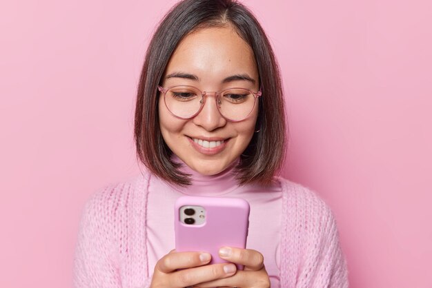 Positive young asian woman holds mobile phone in hands reads\
funny message she received from friend wears optical eyeglasses and\
casual jumper isolated over pink background scrolls newsfeed