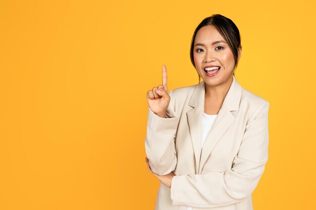 Positive young asian woman in suit point finger up at free space isolated on yellow studio