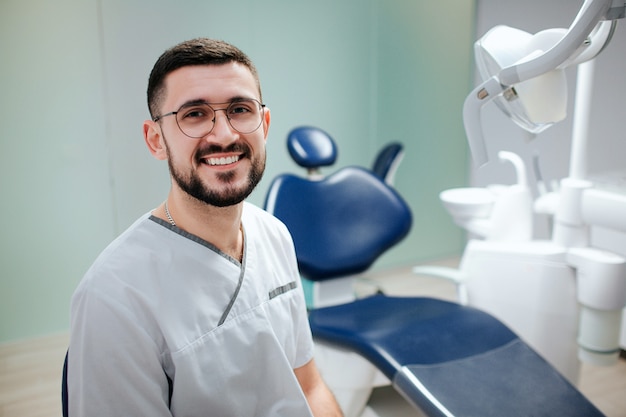 Positive young bearded dentist siting in dentistry room