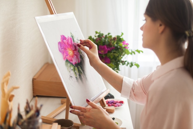 Positive young beautiful smiling girl artist works in studio selects colors of bed for work with pink rosehip flower sitting at table with an easel and a bouquet. Concept creativity as profession