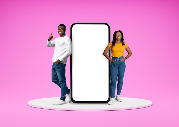 Positive young black couple standing by big smartphone with mockup