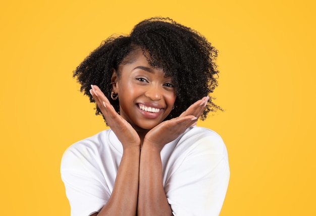 Positive young black curly lady in white tshirt touches face with hands enjoys beauty care