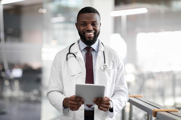 Positive young black doctor holding digital tablet clinic interior