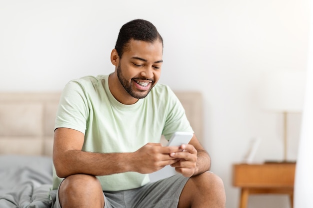 Positive young black guy sitting on bed using cell phone