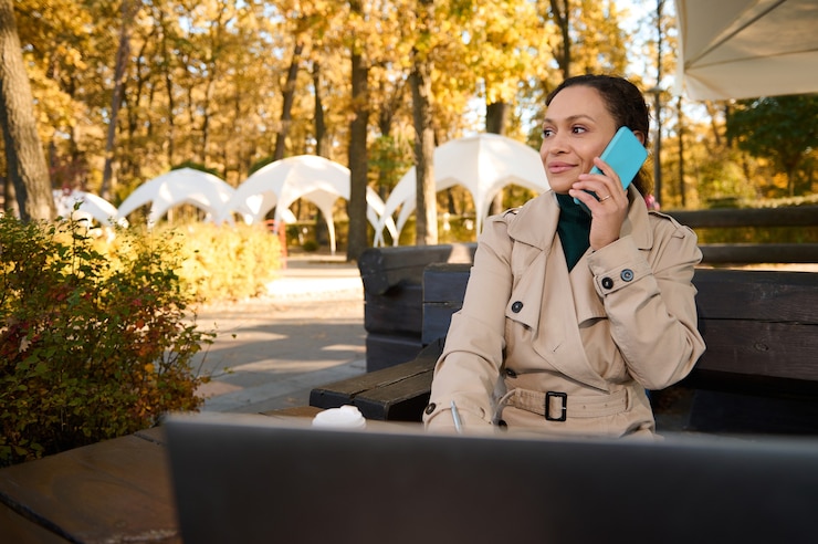 Photo positive young black woman, business person sitting in outdoor cafe and looking away while talking on the mobile phone on the background of oak grove at sunny autumn warm day