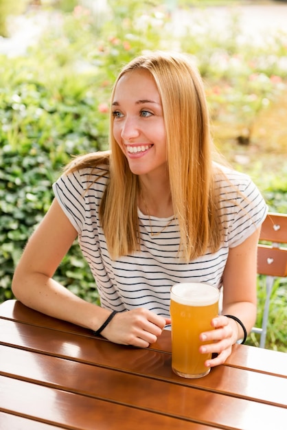 Positive young blond haired female in casual outfit sitting at wooden table with glass full of cold foamy lager beer and smiling while enjoying summer weekend on terrace of restaurant