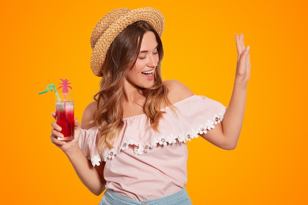 Positive young Caucasian female has fun, wears summer straw hat and blouse, holds summer cocktail, poses against orange