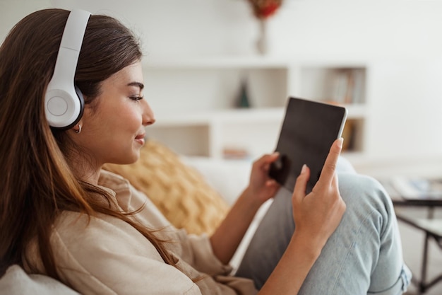 Positive young european woman in wireless headphones watch video on tablet has video call in room