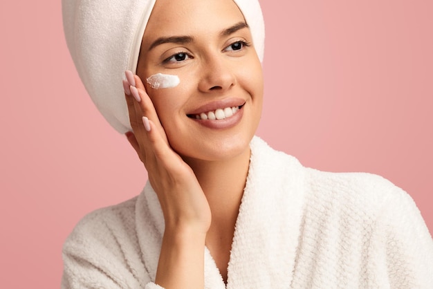 positive young female in bathrobe taking care of her skin applying moisturizing cream on face while on pink background in studio