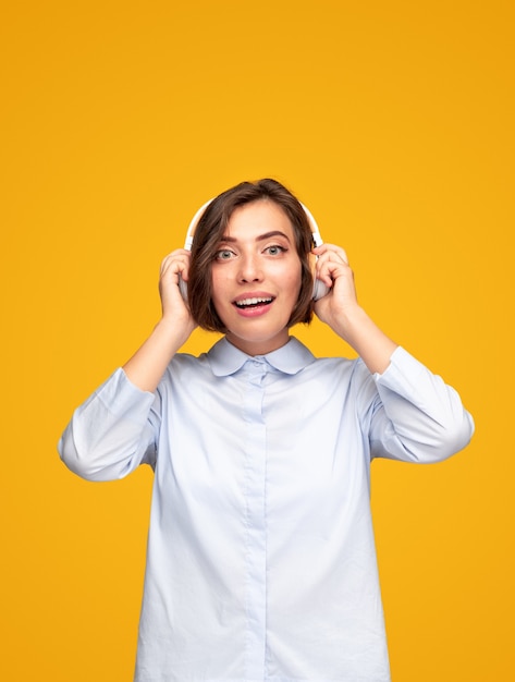 Positive young female in casual shirt looking at camera with excited surprised face expression while listening to music through wireless headphones against yellow background