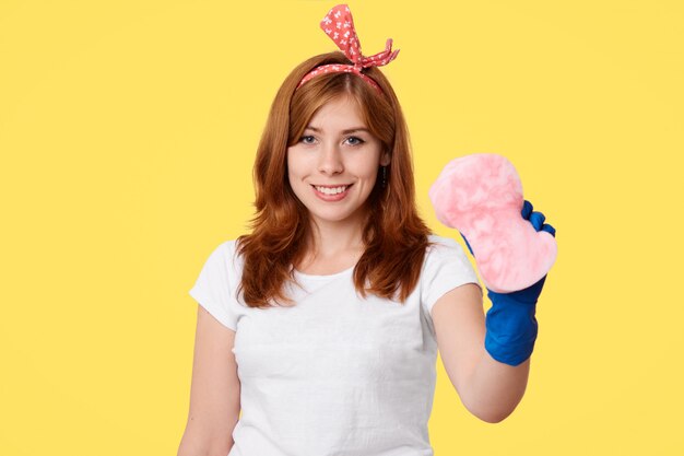 Positive young female does overalls cleaning up in her apartment, enjoys process, wears rubber blue gloves, holds sponge, isolated over yellow wall. Housewife with mop. Cleaning concept