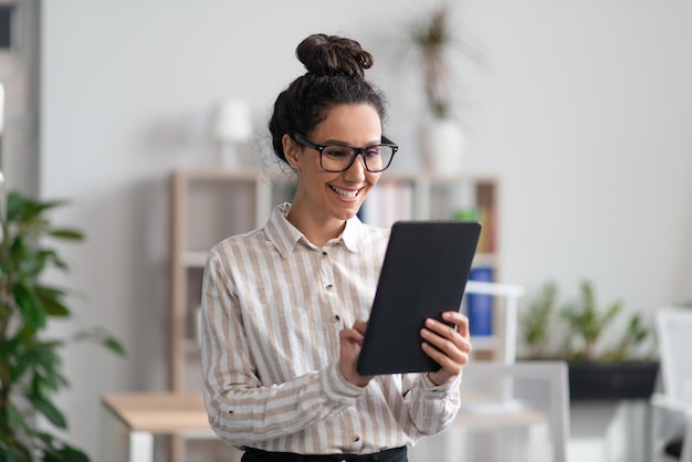 Positive young female entrepreneur using digital tablet in office and smiling checking her agenda on