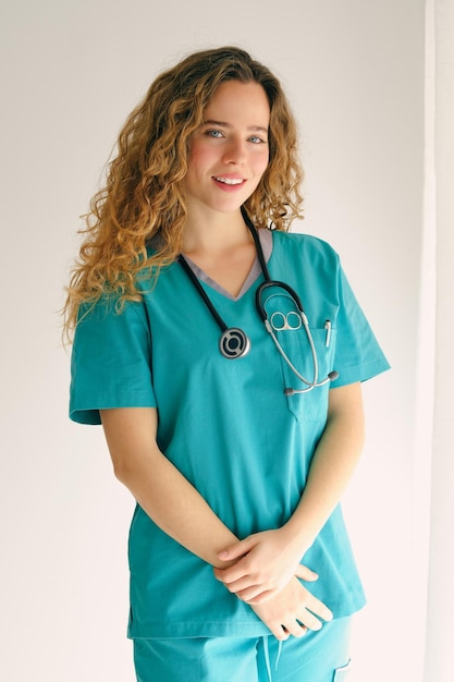 Positive young female physician with long wavy hair in uniform and stethoscope standing near white wall with crossed hands and looking at camera