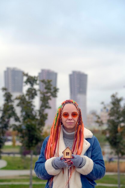 Positive young female in warm clothes and with long dyed dreadlocks holding small wooden house and looking at camera against blurred skyscrapers in city