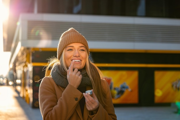 Positive young female with long blond hair in warm coat applying lip balm while standing on street near building during stroll