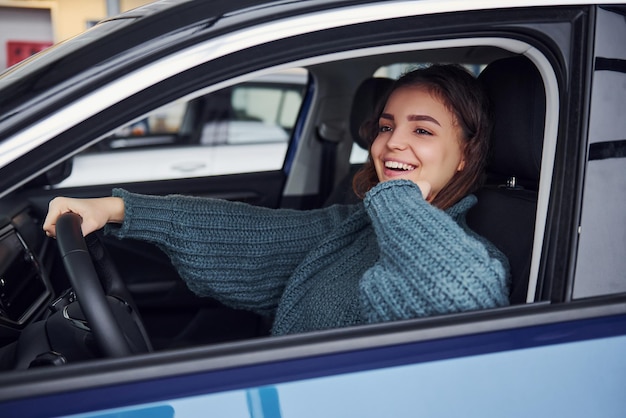 Positive young girl in casual clothes sitting inside of modern car and smiling.