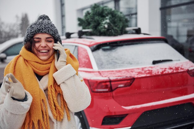 Foto ragazza positiva in piedi vicino all'auto con albero di natale verde in cima.