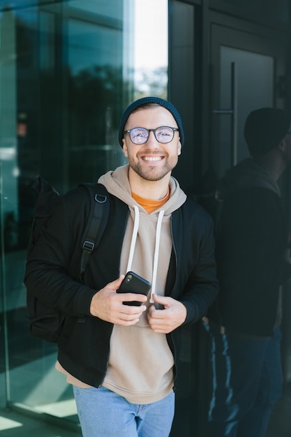 Positive young guy holding smartphone in his hands looking at camera standing on street Bearded man in eyeglass wears casual clothes Cell phone usage concept