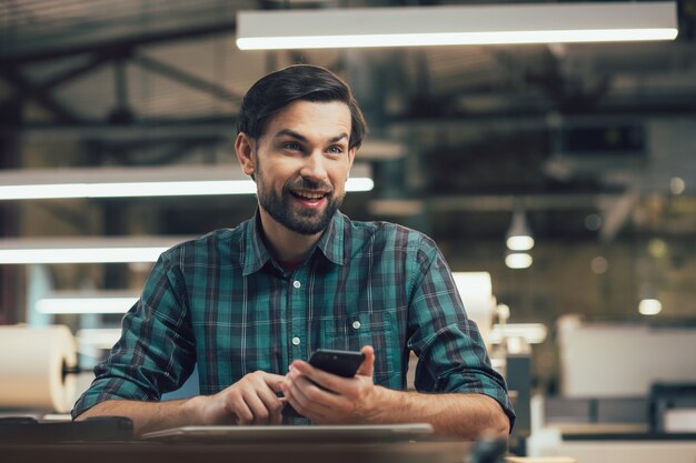 Positive young man in checkered shirt sitting at the table and holding a smartphone