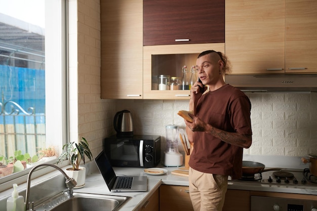 Positive young man eating sandwich for breakfast in kitchen and talking on phone with coworker or fr