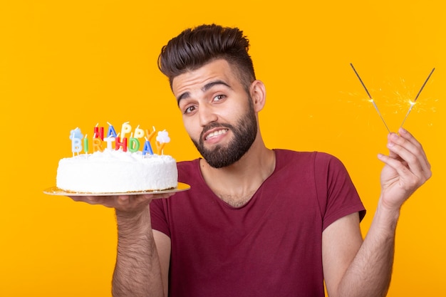 Positive young man holding a happy birthday cake and two burning bengal lights posing on a yellow surface. Advertising space