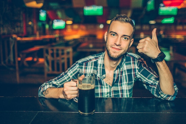 Positive young man sit at bar counter in pub and look . He hold big thumb up. Guy has hand on mug with dark beer.