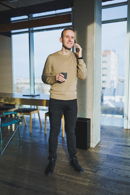 Позитивный молодой человек улыбается и разговаривает по мобильному телефону и пьет кофе, отдыхая, стоя в офисе Обеденный перерыв во время работы